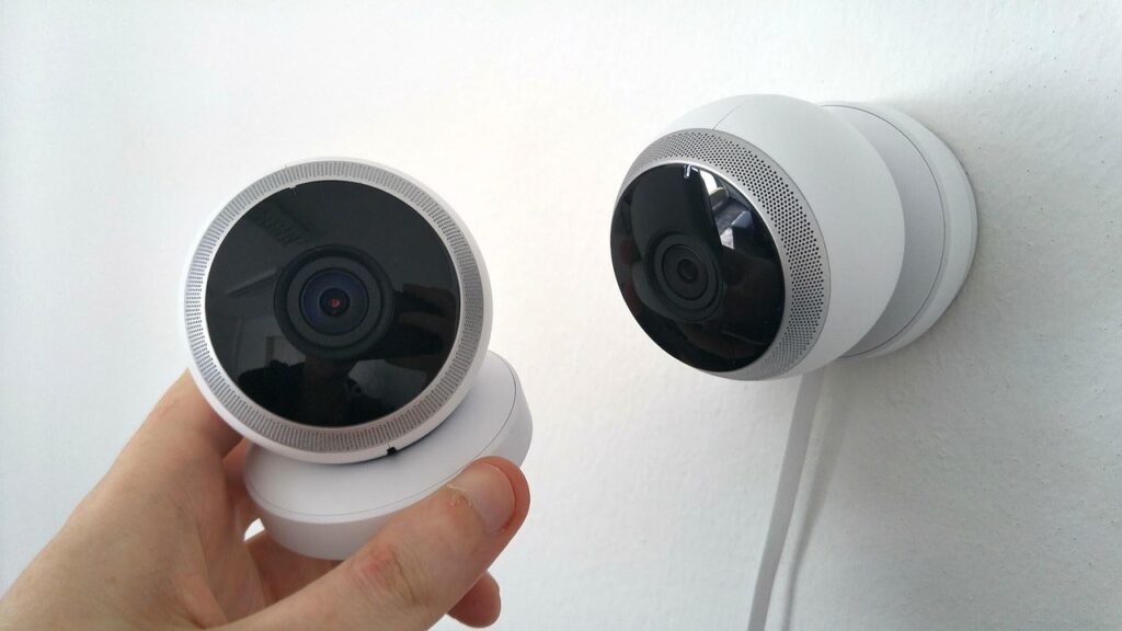 A hand holds a round security camera next to another identical camera mounted on a white wall, showcasing sleek consumer electronics manufacturing.