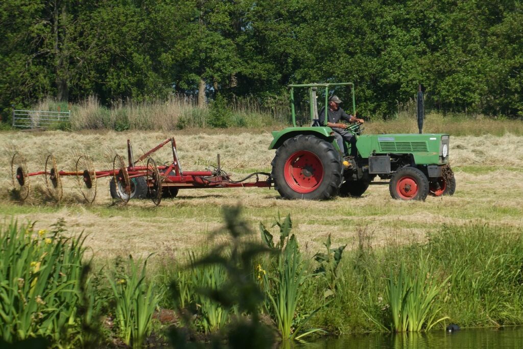 A person drives a green tractor equipped with agricultural electronics, pulling a red hay rake through a field of cut grass, with trees and tall plants in the background.