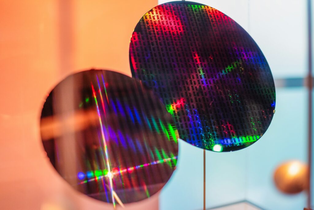 Two silicon wafers displaying colourful light reflections, set against a blurred, warm-toned background, highlight the precision of semiconductor electronics manufacturing.