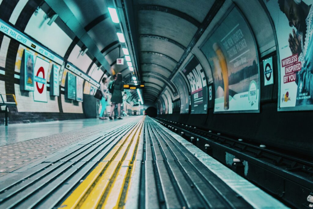 Low-angle view of a London Underground platform with yellow tactile paving, advertising posters on tunnel walls, and a few people waiting in the distance—showcasing how rail electronics manufacturing supports modern transport systems.