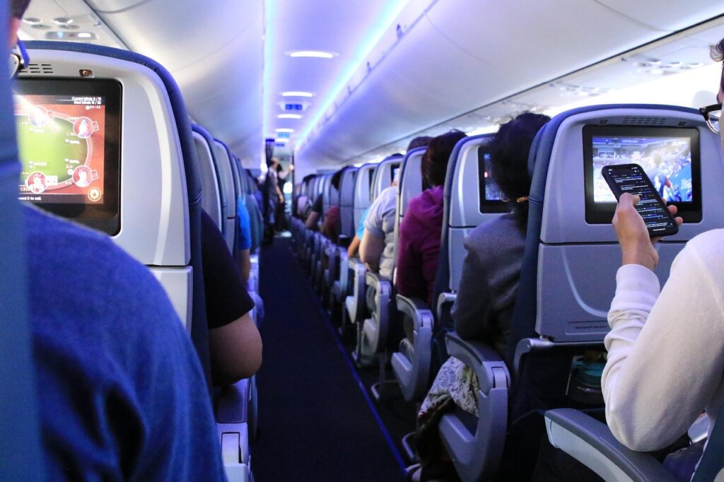 Passengers seated inside an aeroplane cabin, some using in-flight entertainment screens and mobile devices—innovations made possible by advancements in aerospace electronics manufacturing, with overhead lights illuminating the scene.