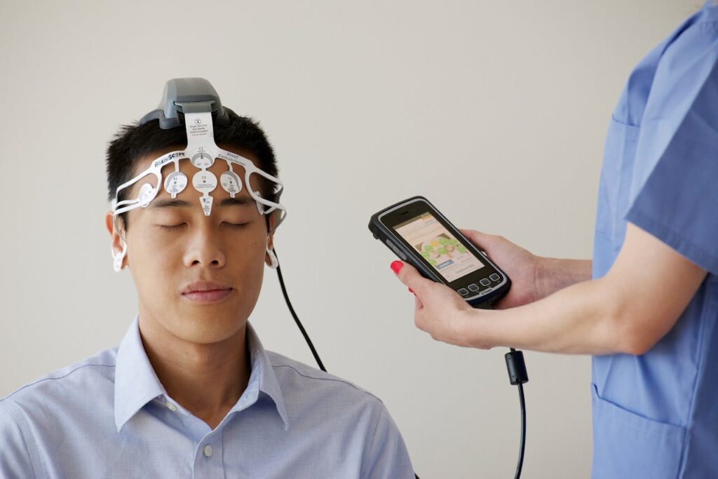 A person sits with eyes closed wearing a head-mounted device, while another in scrubs—reflecting the advancements of medical electronics manufacturing—holds a handheld monitor displaying data.