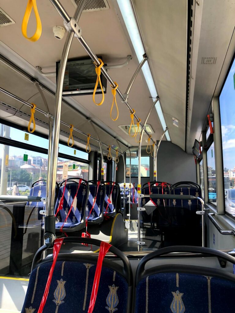 The interior of a city bus with blue patterned seats, yellow hand straps, and red markings indicating some seats are unavailable highlights advances in automotive electronics manufacturing. Daylight streams through the windows.