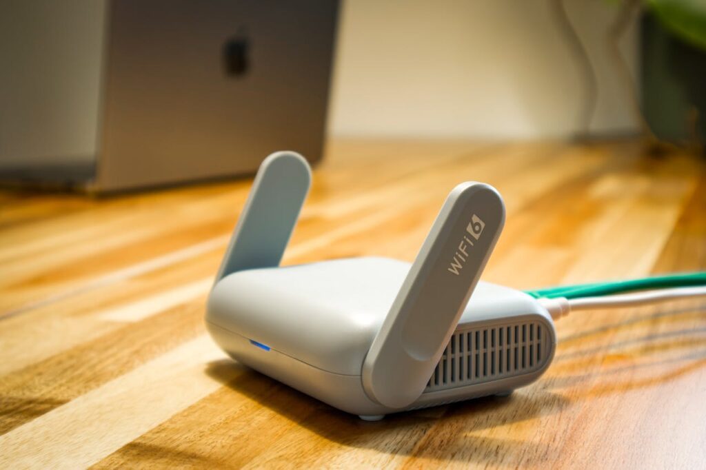 A WiFi 6 router from a leading telecoms electronics manufacturing brand sits on a wooden desk, connected by a green Ethernet cable, with a laptop in the background.
