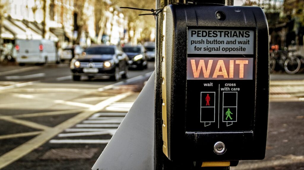 Close-up of a pedestrian crossing signal with the word "WAIT" lit up, featuring advanced automotive electronics manufacturing, mounted at a zebra crossing with cars and buildings visible in the background.