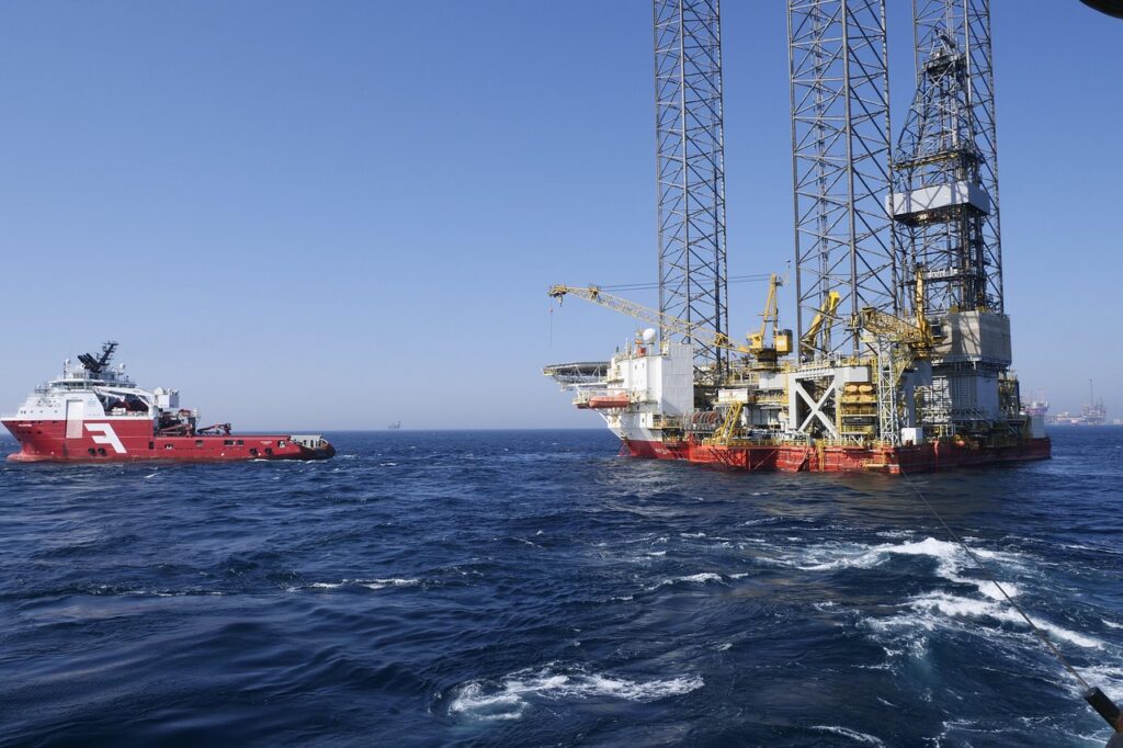 A red supply vessel approaches an offshore oil and gas rig with tall lattice towers in the open blue sea under a clear sky.