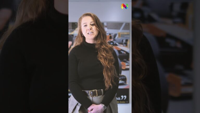 A woman with long, wavy red hair stands indoors, wearing a black polo neck and grey trousers with a belt. A blurred background hints at an electronics manufacturer, featuring car images and a colourful logo in the top right.