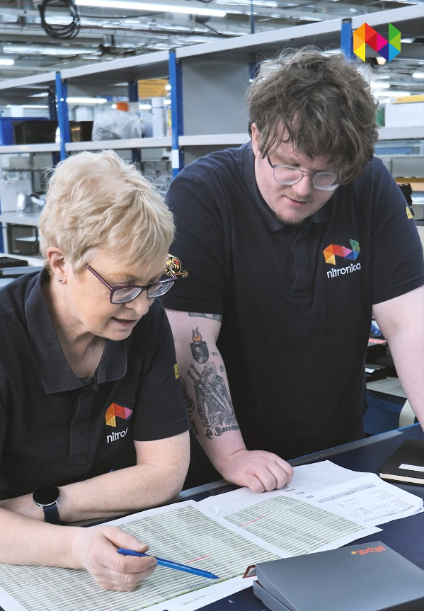Two people in matching company uniforms review paperwork together at a workstation in a Contract Electronics Manufacturing Services warehouse or factory setting.